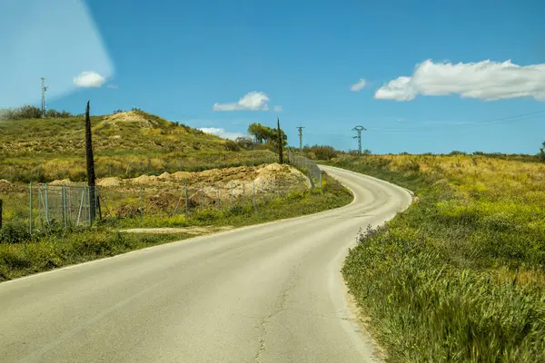 Kırsal alanda dolambaçlı bir yol, parlak mavi bir gökyüzü ve kabarık bulutların altında yeşilliklerle doludur.