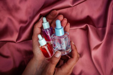 Kozmetik şişeleri açık kadın ellerinde narin pembe saten kumaş üzerinde dururlar. Sıvı ve saten renkler sakinleştirici estetik yaratır.