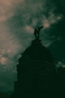 Madrid, İspanya 1 Mayıs 2024 Bir melek heykeli ile bir anıt silueti çarpıcı bir kontrast yaratan bulutlu bir gökyüzü karşısında dramatik bir şekilde duruyor