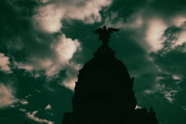 Madrid, İspanya 1 Mayıs 2024 Bir melek heykeli ile bir anıt silueti çarpıcı bir kontrast yaratan bulutlu bir gökyüzü karşısında dramatik bir şekilde duruyor