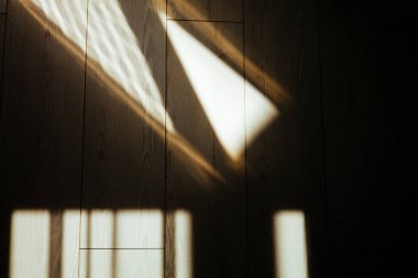 Güneş ışığı, ahşap zeminde geometrik gölgeler yaratarak soyut desenler oluşturur. Ahşap zemin ve pencere ve gölgeleri gösteren soyut resim.