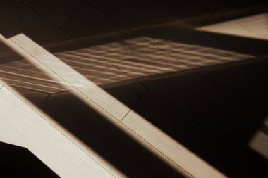 Güneş ışığı, ahşap zeminde geometrik gölgeler yaratarak soyut desenler oluşturur. Ahşap zemin ve pencere ve gölgeleri gösteren soyut resim.