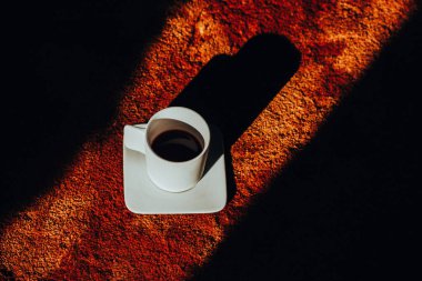 Bir fincan tabağında beyaz bir kahve fincanı parlak ışıkla çelişen dokulu kahverengi bir halının üzerinde uzun bir gölge bırakır. Rahat evde sabah içkisi..