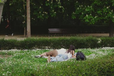 Madrid, İspanya 1 Mayıs 2024 Retiro parkında bir çift, yeşilliklerle çevrili bir battaniyenin üzerinde dinleniyor. Durgun bir sahne, bir bank ve çiçek açan doğa..