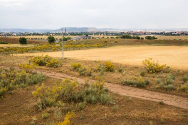 Geniş bir manzara, sergilenen tarlalar, toprak yollar, ve bulutlu bir gökyüzü altında uzak bir kasaba, bir huzur hissi uyandırıyor. Sonbahar doğası.