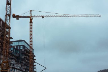 Bulutlu bir gökyüzünün altında kısmen inşa edilmiş bir binanın yanında yüksek bir vinç yükseliyor. İnşaat alanında kentsel gelişim sergileniyor. Emlak.