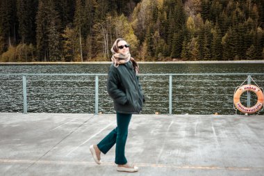 Doğayla güzel bir günün tadını çıkaran neşeli bir kadın dingin göl kenarındaki rıhtımda yürüyor. Güneş gözlüğü ve eşarp takmış gülümseyen bir kız.. 