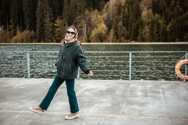 Doğayla güzel bir günün tadını çıkaran neşeli bir kadın dingin göl kenarındaki rıhtımda yürüyor. Güneş gözlüğü ve eşarp takmış gülümseyen bir kız.. 