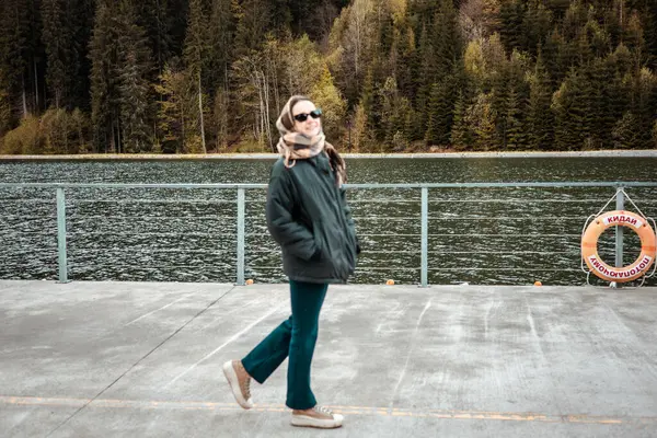 Doğayla güzel bir günün tadını çıkaran neşeli bir kadın dingin göl kenarındaki rıhtımda yürüyor. Güneş gözlüğü ve eşarp takmış gülümseyen bir kız.. 