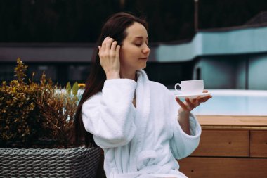Rahat bir kadın, huzurlu bir ortamda beyaz bir bornoz giyerek havuz kenarında çay içmekten zevk alır. Huzurlu bir kız sabah kahvesini yudumlarken eğlenir..