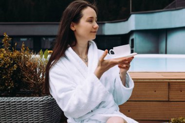 Rahat bir kadın, huzurlu bir ortamda beyaz bir bornoz giyerek havuz kenarında çay içmekten zevk alır. Huzurlu bir kız sabah kahvesini yudumlarken eğlenir..