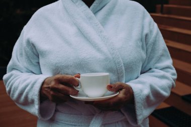 Beyaz bornoz giyen rahat bir kadın, elinde bir fincan kahveyle huzurlu bir anın tadını çıkarıyor. Yumuşak bornozlu bir adam elinde kupa, otelde dinleniyor..