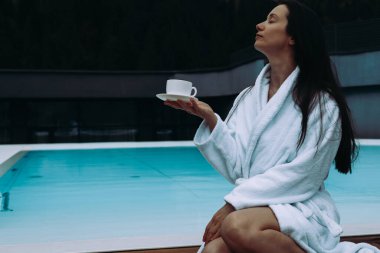 Rahat bir kadın havuz kenarında beyaz bir bornoz giyerek çay içmekten hoşlanır. Beyaz cüppeli sakin bir bayan spa otelinde bir fincan çayla eğleniyor. Beyaz cüppeli sakin bir kadın bir fincan çayla sakin bir havuzun yanında dinleniyor.