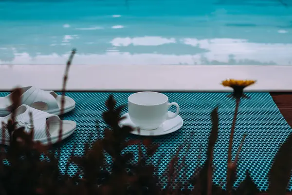 Sakin bir sahnede yazın havuz kenarında bir fincan, terlik ve çiçek sergilenir. Sakin bir havuz kenarında içki içilir. Sakin mavi su. Yaz havası. 