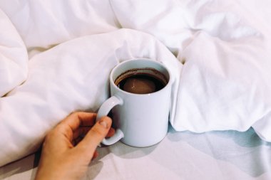 Bir insanın eli, bir yatağın yumuşak beyaz çarşaflarına yerleştirilmiş dumanı tüten bir fincan kahveye uzanır. Basit bir sahne samimi ve davetkar bir atmosfer yaratır.