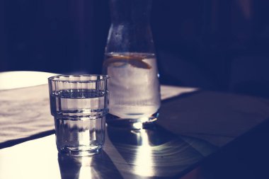 Bir bardak su ve limonlu bir sürahi. Limon dilimli ferahlatıcı bir su loş bir restoran ortamında sakin bir sahne yaratıyor. Tablo ayarı