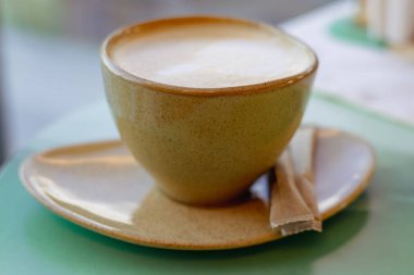 Kahverengi bir fincanda, rahatlatıcı bir kahve molası ya da sabah ayinine hazır güzel sunulmuş bir kapuçino. Sütlü sıcak, davetkar bir fincan kahve, şeker paketleriyle dolu bir fincan tabağında, yumuşak bir odak ile oturur..