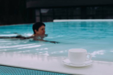 Bir insan havuzda bir fincan kahveyle birlikte yüzer, bir dakikalık rahatlığın tadını çıkarır beyaz bir fincan ve çay tabağı havuzun kenarında otururken, bir insan huzur ve sükunet anı yakalar.