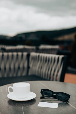 Kahve fincanı ve dışarıda mermer bir masada şık bir güneş gözlüğü ile huzurlu bir ortam. Beyaz bir fincan ve siyah bardaklar mermer bir masada otururlar. Rahatlatıcı bir gün. Güzel bulutlu bir manzara sahneyi tamamlıyor.