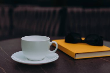 Masasında siyah güneş gözlüğü olan beyaz bir fincan ve sarı bir defter. El değmemiş beyaz bir fincan sarı bir kitabın yanında oturur, rahatlamak ve okumak için mükemmeldir..