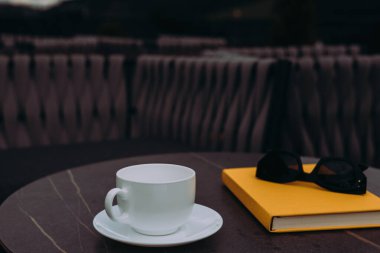 Masasında siyah güneş gözlüğü olan beyaz bir fincan ve sarı bir defter. El değmemiş beyaz bir fincan sarı bir kitabın yanında oturur, rahatlamak ve okumak için mükemmeldir..