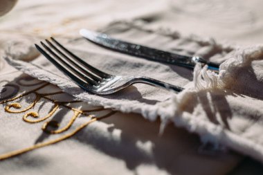 Parlak bir çatal ve bıçak kumaşlı bir masa örtüsünün üzerinde dinleniyor, yemek için hazır. Bej masa örtüsüne yakın plan bir çatal bıçak seti. Yemek sahnesi
