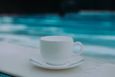 Beyaz bir çay fincanı ve çay tabağı bulanık mavi su arka planlı bir havuzun yanında oturur. Görüntü, rahatlama ve rahatlama dolu sakin bir atmosferi çağrıştırıyor..