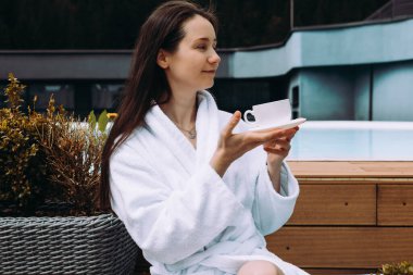 Rahat bir kadın, huzurlu bir ortamda beyaz bir bornoz giyerek havuz kenarında çay içmekten zevk alır. Huzurlu bir kız sabah kahvesini yudumlarken eğlenir..