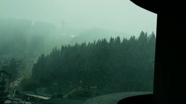 Dağlarda kış fırtınası altında ağaçlarla, dağlarla ve köyle kaplı karlı bir manzara. Bir kış fırtınası sırasında ağaçlarla, dağlarla ve uzak bir köyle karlı bir arazinin manzarası..