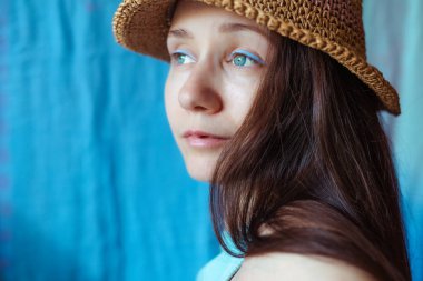 Mavi göz kalemi olan sakin bir genç kadın hasır şapka takıyor ve mavi bir fona karşı düşünceli bir ifadeyle uzaklara bakıyor..