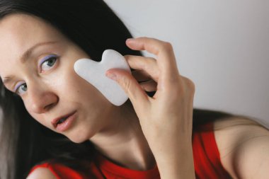Kadın cildini geliştirmek için yüzüne gua sha aleti ile nazikçe masaj yapıyor. Yüzünde gua sha aleti kullanan bir kadın. Yüz masajı yaparken aleti parmaklarıyla tutuyor..