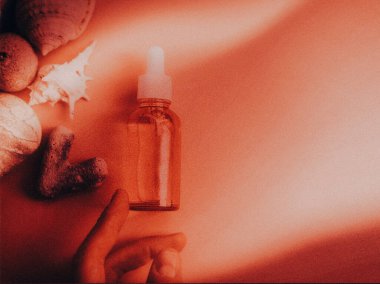 Sıcak bir zemin üzerinde deniz kabuğu ve mercan bulunan cilt serumu şişesi. Bir el, sıcak ve desenli bir arka planda deniz kabuğu ve mercanla çevrili bir şişeye doğru uzanır. Boşluğu kopyala.