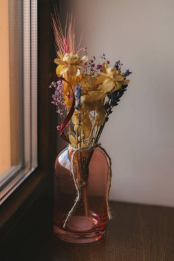 Pencerenin yanındaki pembe vazoda kurumuş çiçeklerin güzel bir gösterisi. Bir pencerenin yanında duran gül rengi cam vazoda kurumuş, narin bir çiçek aranjmanı, huzurlu ve rahat bir atmosfer yaratıyor..
