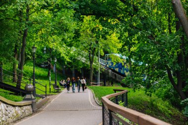 Kyiv, Ukrayna 14 Haziran 2025 İnsanlar parkta ağaçlarla kaplı güzel bir patika boyunca yürümekten zevk alıyor. Ağaçlı sakin bir park manzarası, bir füniküse götürüyor, ve insanlar rahatça geziniyor..