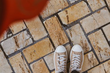 Tuğla kaldırımda bir çift bej spor ayakkabı, sıradan ve şık bir sahne. Sol üst köşesinde kırmızı aksanlı, tuğladan yapılmış kaldırımda duran bej renkli spor ayakkabıları..