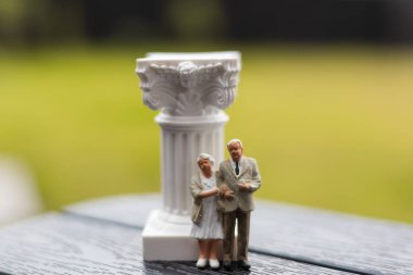 Minyatür yaşlı bir çift dekoratif beyaz bir sütunun yanında poz veriyor. Yaşlı insanlar - emekli bir adam ve bir kadın - yumuşak, bulanık yeşil bir arka planı olan dekoratif bir sütunun yanında duruyor. Emeklilik planı