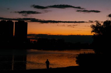 Canlı günbatımı gölün üzerinde sıcak bir parıltı ve yalnız bir balıkçı bırakıyor. Çarpıcı bir günbatımı, sakin bir göl, siluetli binalar ve balık tutan bir kişi, sakin ve huzurlu bir atmosfer yaratıyor..