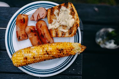 Beyaz bir tabakta ızgara sosis, mısır ve ekmek. Lezzetli bir tabak ızgara yemek, yaz barbeküsü için mükemmel. İçinde sosis, mısır ve ekmek var..