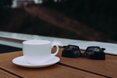 Dışarıdaki ahşap masada güneş gözlüklü bir fincan kahve. Beyaz çay fincanı ve çay tabağı, arka planda bulanık güneş gözlüklerinin yanında, ahşap bir yüzeyde oturur..