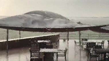 Yağmurlu bir havada bir kafede ya da restoran terasında şemsiyeyle ya da koruyucu bir gölgelikle ıslak bir pencereden bakın. Yağmurlu bir günde pencereye yağmur damlaları, masalar ve sandalyeler. Şiddetli rüzgar. 