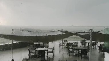 Yağmurlu bir havada bir kafede ya da restoran terasında şemsiyeyle ya da koruyucu bir gölgelikle ıslak bir pencereden bakın. Yağmurlu bir günde pencereye yağmur damlaları, masalar ve sandalyeler. Şiddetli rüzgar. 