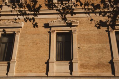 Ayrıntılı pencereler ve ağaç dalları altında dekoratif süslemelerin yer aldığı güneş ışığıyla aydınlatılmış mimari ön cephe. Pencereli, güneş ışığıyla aydınlatılmış, eski bir evin mimari görüntüsü. Detaylı süslemeler.