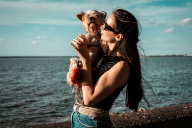 Bir kadın ve Yorkshire teriyeri güneşli bir günde suyun kenarında tatlı bir anı paylaşıyorlar. Bir kız suyun kenarında küçük bir köpeği kucaklıyor, dışarıda sevgi ve arkadaşlığın tadını çıkarıyor. Hayvan sahibi.