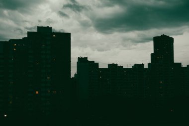 Gece dramatik bir şehir manzarasında karanlık bulutlu kasvetli gökyüzüne karşı binaların siluetleri yer alır. Slhouetted bölge bulutlu ufuk çizgisi altında