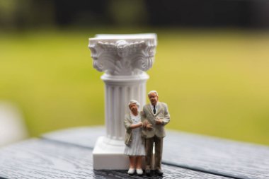 Minyatür yaşlı bir çift dekoratif beyaz bir sütunun yanında poz veriyor. Yaşlı insanlar - emekli bir adam ve bir kadın - yumuşak, bulanık yeşil bir arka planı olan dekoratif bir sütunun yanında duruyor. Emeklilik planı