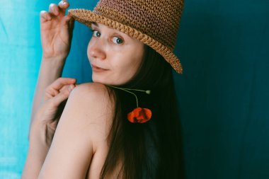 Çekici genç kadın elinde kırmızı bir gelincik çiçeğiyle hasır şapkası ile poz veriyor. Güneş şapkalı bir kız, neşeli bir bakışla omzunun üzerinden bakıyor.