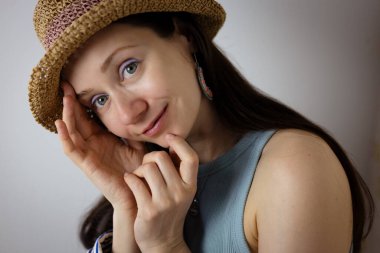 Hasır şapkalı bir kadın gülümsüyor, yumuşak ışıklandırmalı davetkar bir portre oluşturuyor. Şapka, kolye ve halka küpeler takan bir kadın gülümseyerek doğal ışıkta sıcak, davetkar bir portre çizer..