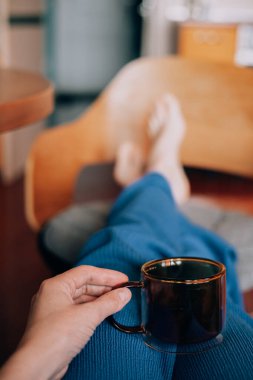 Evde bir fincan kahveyle rahatlamak, huzurlu bir sabah geçirmek bir kupa çayla dinlenmek, ayaklar sandalyede, ev ortamında rahatlamak. Yüzü olmayan bir kadın evdeki koltukta oturuyor..