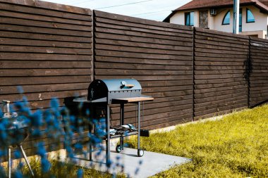 Bir barbekü ızgarası ve avlu arka bahçede yemek pişirmek için hazır. Arka bahçedeki ahşap bir çitin önünde barbekü ızgarası. Arka bahçede çimenler ve arka planda bir ev var. Hafta sonu tatili