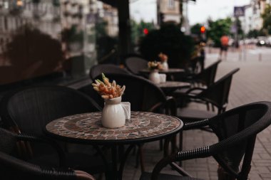 Masaları, sandalyeleri ve bir vazo kurutulmuş çiçekleri olan büyüleyici bir kafe terası müşterileri bekliyor. Dekoratif masaları ve sandalyeleri olan açık hava kafe sahnesi müşteriler için hazır..